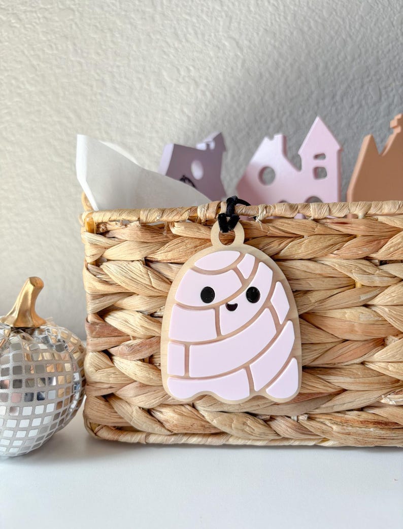 May include: A light-colored, ghost-shaped pendant with a pink design and black eyes and mouth hangs from a black cord. The pendant is attached to a woven basket. A silver disco ball pumpkin is to the left.