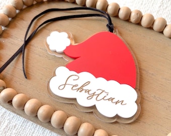 May include: A red Santa hat-shaped Christmas ornament with white trim and the name "Sebastian" in a cursive font. The ornament hangs from a black string and is set on a wooden tray with wooden beads.