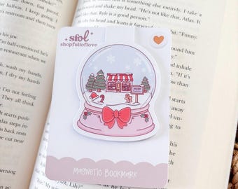 May include: A pink magnetic bookmark featuring a snow globe design with a shop, trees, and snowflakes. The bookmark has the text "shopfulloflove" and "MAGNETIC BOOKMARK". The bookmark is placed on an open book.