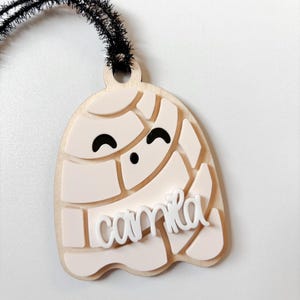 May include: A light-colored, layered ghost-shaped ornament with the name "Camila" in white script. The ghost has a smiling face and a black fuzzy wire for hanging. The ornament is likely made of wood and acrylic.