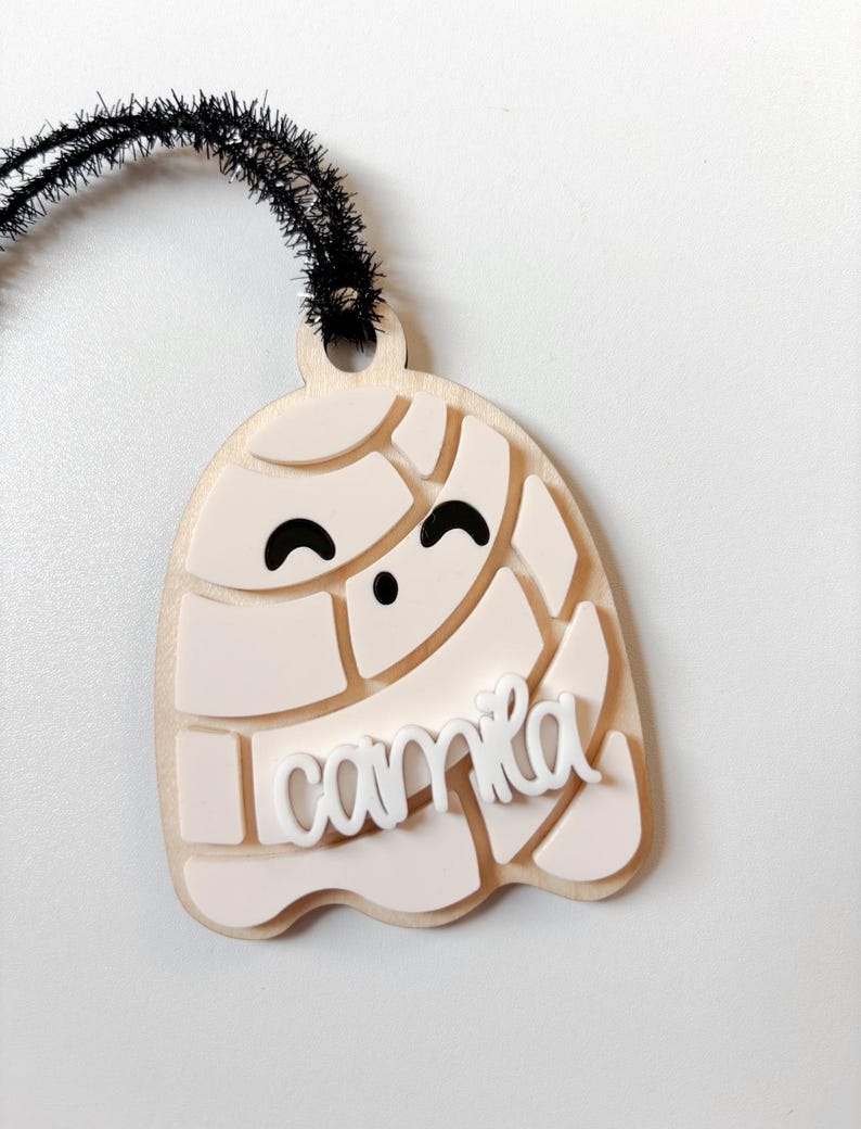 May include: A light-colored, layered ghost-shaped ornament with the name "Camila" in white script. The ghost has a smiling face and a black fuzzy wire for hanging. The ornament is likely made of wood and acrylic.