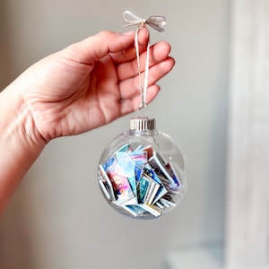 May include: A clear glass Christmas ornament filled with colorful miniature books. The ornament has a silver top and a white ribbon with gold accents tied around it.