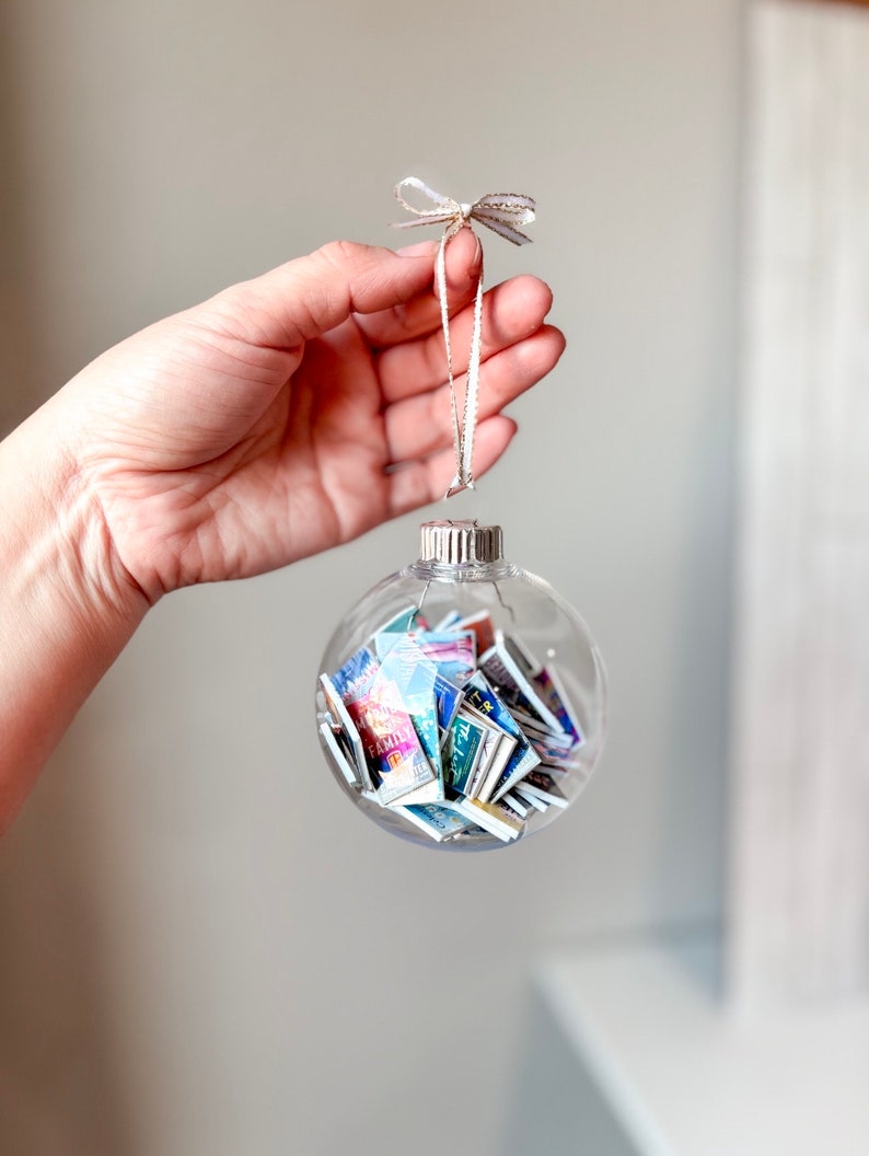 May include: A clear glass Christmas ornament filled with colorful miniature books. The ornament has a silver top and a white ribbon with gold accents tied around it.
