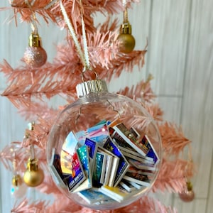 May include: A clear glass Christmas ornament filled with miniature book covers. The ornament is hanging on a pink artificial Christmas tree.