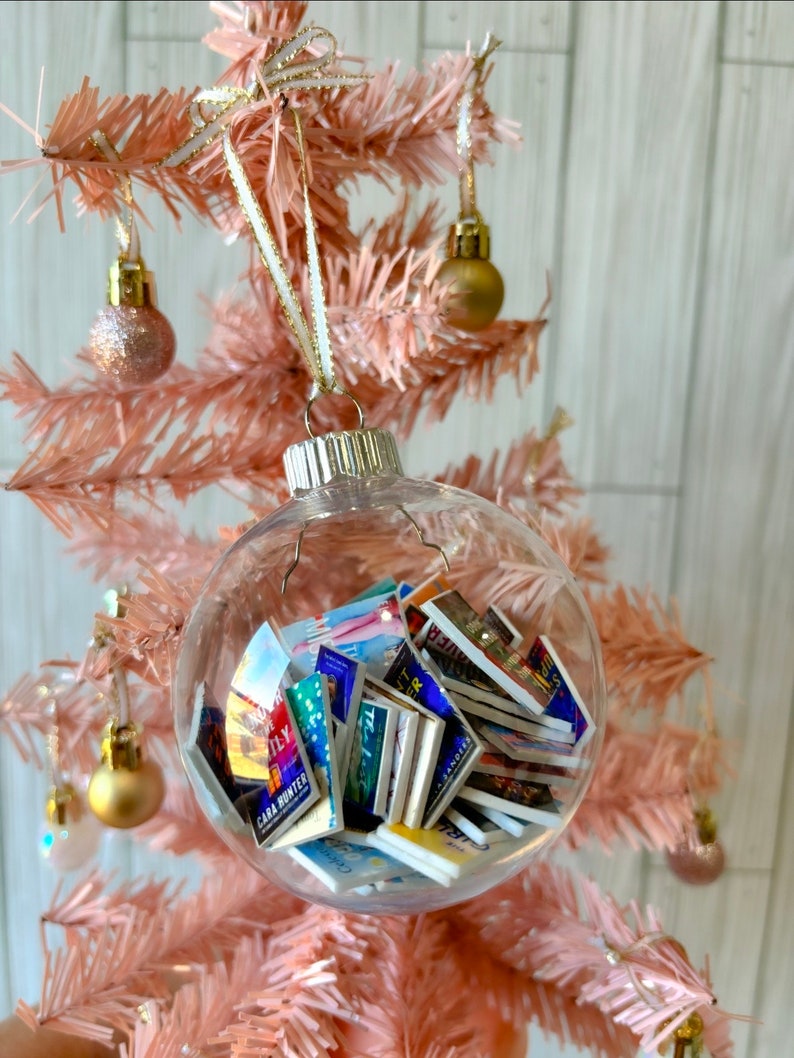 May include: A clear glass Christmas ornament filled with miniature book covers. The ornament is hanging on a pink artificial Christmas tree.
