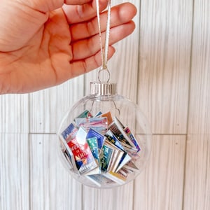 May include: A clear glass ornament filled with miniature book covers. The ornament is hanging from a gold and white ribbon.