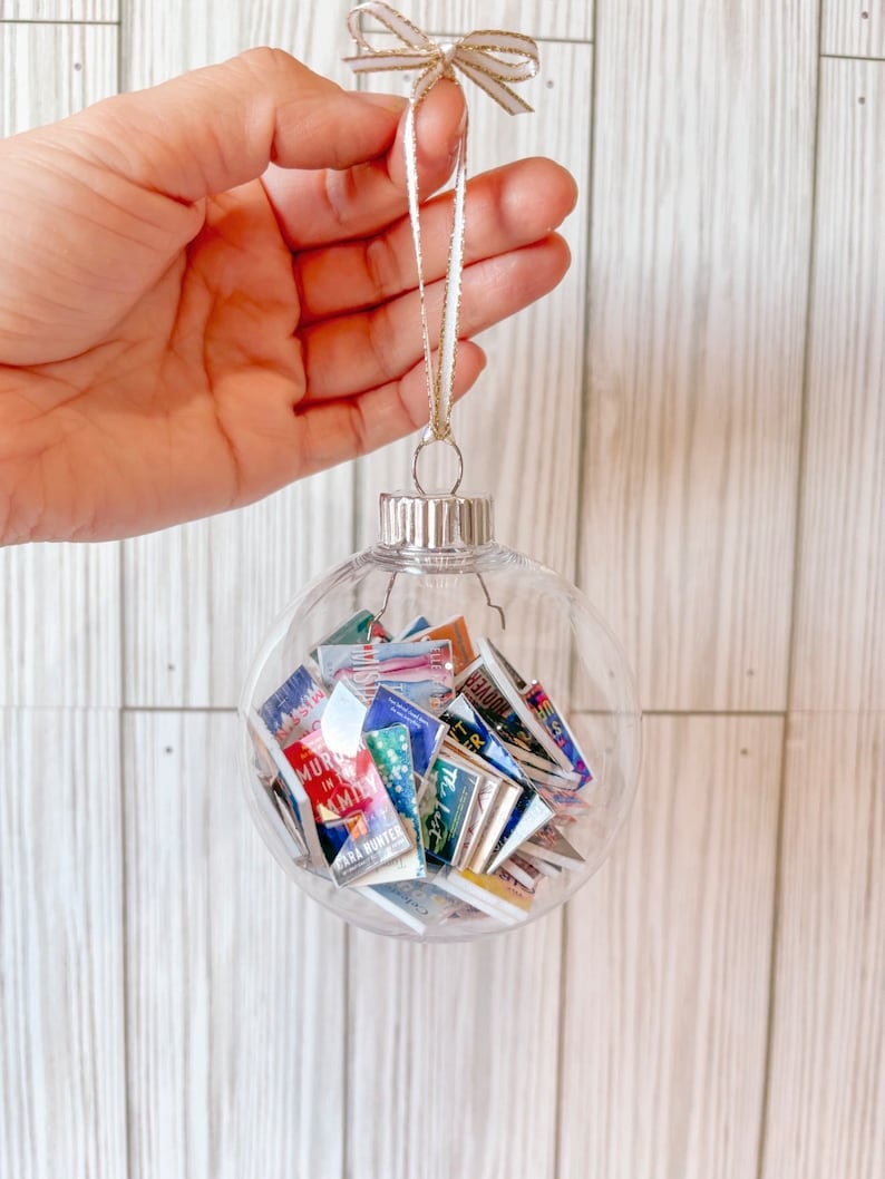 May include: A clear glass ornament filled with miniature book covers. The ornament is hanging from a gold and white ribbon.