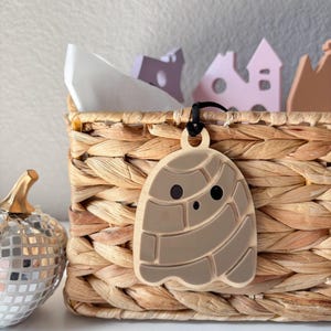 May include: A light brown, concha-shaped pendant with a black cord hangs in front of a woven basket. The pendant has a smiling face and a detailed design. A silver disco pumpkin and small wooden houses are also visible.