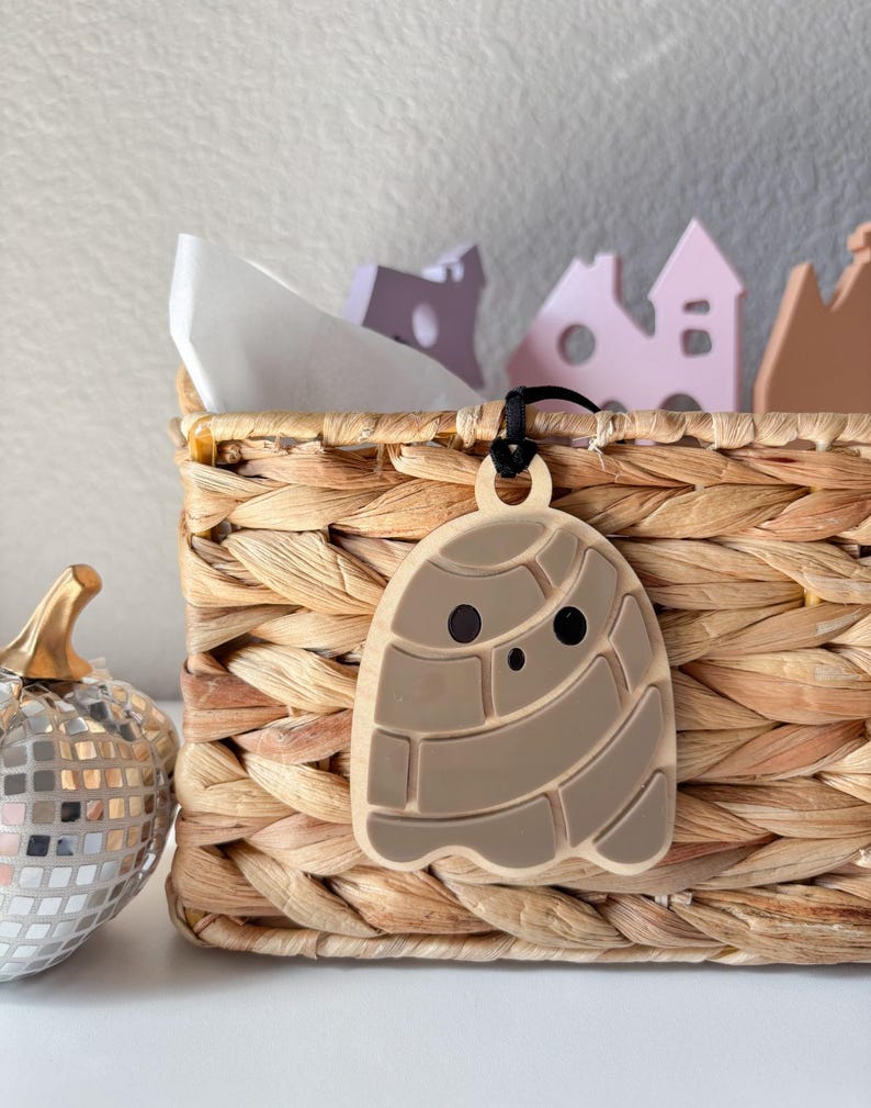 May include: A light brown, concha-shaped pendant with a black cord hangs in front of a woven basket. The pendant has a smiling face and a detailed design. A silver disco pumpkin and small wooden houses are also visible.