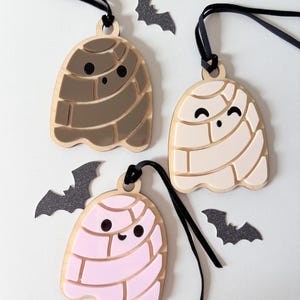 May include: Three wooden ghost ornaments in brown, cream, and pink, each with a black string for hanging. The ghosts have a smiling face and a segmented design. Black bat cutouts are scattered around the ornaments, creating a Halloween theme.