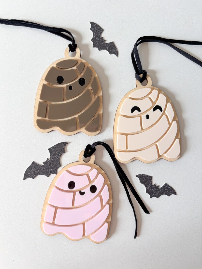 May include: Three wooden ghost ornaments in brown, cream, and pink, each with a black string for hanging. The ghosts have a smiling face and a segmented design. Black bat cutouts are scattered around the ornaments, creating a Halloween theme.
