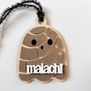 May include: A wooden ghost-shaped ornament with the name "malachi" in white block letters. The ghost has a light brown base with darker brown segmented details and black dot eyes. A black fuzzy wire is attached at the top.