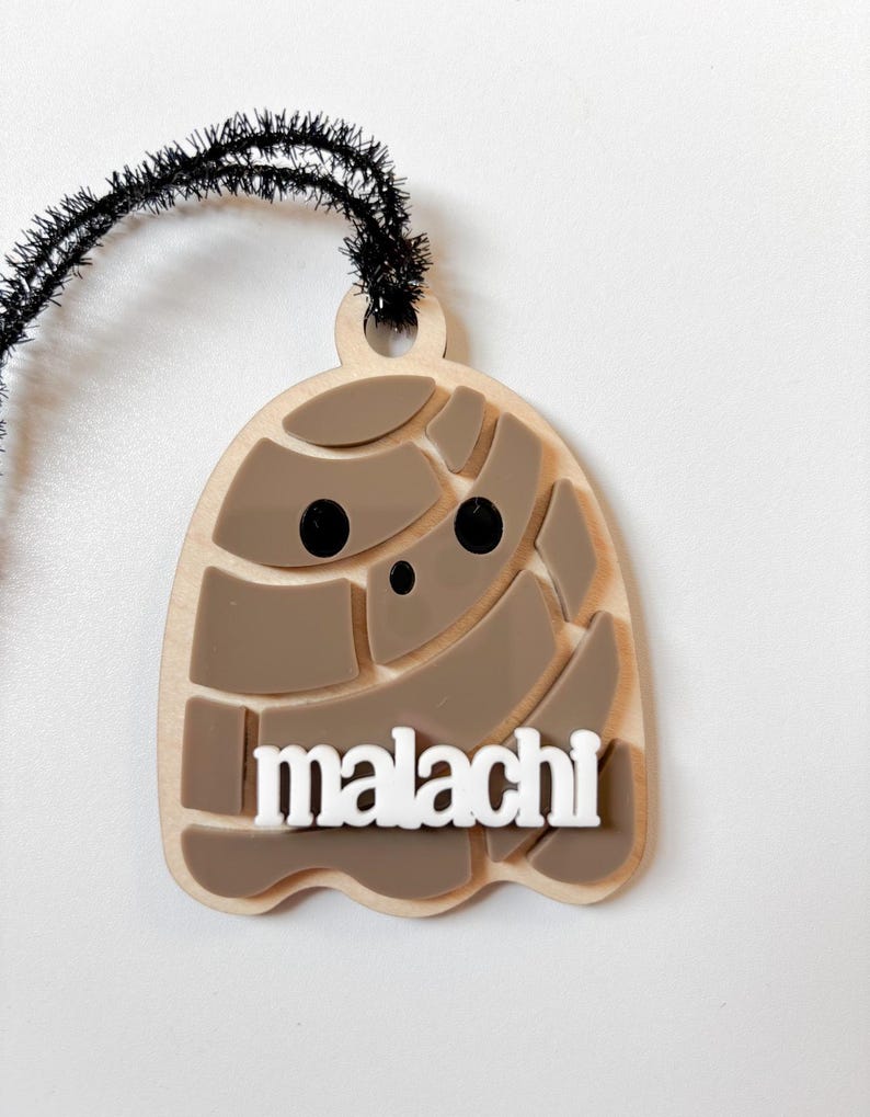 May include: A wooden ghost-shaped ornament with the name "malachi" in white block letters. The ghost has a light brown base with darker brown segmented details and black dot eyes. A black fuzzy wire is attached at the top.
