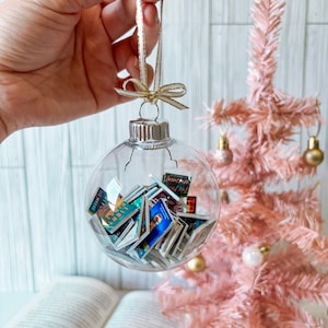 May include: A clear glass Christmas ornament filled with miniature book covers. The ornament is hanging from a white ribbon with a gold bow. The ornament is in front of a pink artificial Christmas tree.