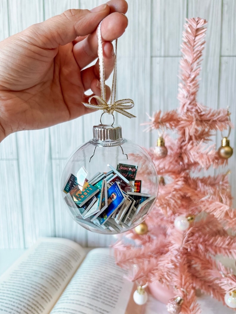 May include: A clear glass Christmas ornament filled with miniature book covers. The ornament is hanging from a white ribbon with a gold bow. The ornament is in front of a pink artificial Christmas tree.