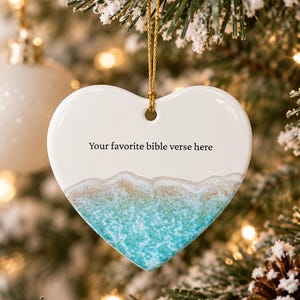 Può includere: Un ornamento in ceramica bianca a forma di cuore appeso a un cordino dorato. La parte inferiore presenta un disegno stampato di una spiaggia sabbiosa e onde del mare turchese. Il testo "Your favorite bible verse here" è stampato in nero.
