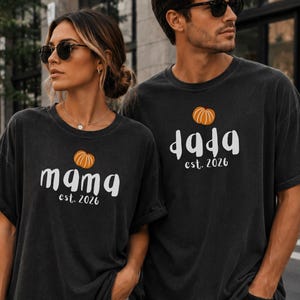 May include: Two charcoal grey short-sleeve t-shirts featuring a small orange pumpkin graphic above white text. One shirt displays the word mama and est. 2026, while the other shirt displays the word dada and est. 2026 in a rounded, casual font style.