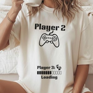 May include: Off-white t-shirt featuring the text "Player 2" and a controller graphic. Below, "Player 3: Loading" is printed with a loading bar and footprints. The shirt is a casual, unisex style.