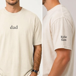 Może przedstawiać: Kremowa koszulka z napisem "dad" wydrukowanym na czarno na piersi. Na rękawie widnieje imię "Kylie Sam" w kolorze czarnym. Koszulka jest w stylu casual, z krótkim rękawem.
