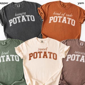 May include: Five short-sleeve crewneck t-shirts in charcoal, orange, olive green, cream, and brown. Each shirt features the word POTATO in a collegiate font with a descriptive adjective above it: hungry, kind of rude, feral, sweet, and sarcastic.