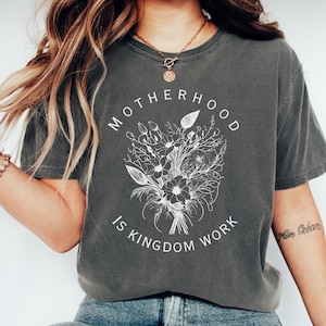 May include: A dark gray t-shirt with a white graphic design that says "Motherhood is Kingdom Work" with a floral illustration.