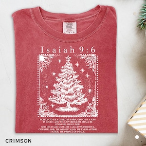 Puede incluir: Camiseta de color carmesí con el texto "Isaías 9:6" y un diseño de árbol de Navidad blanco. La camiseta también presenta una cita de Isaías. La prenda está hecha de un material suave.