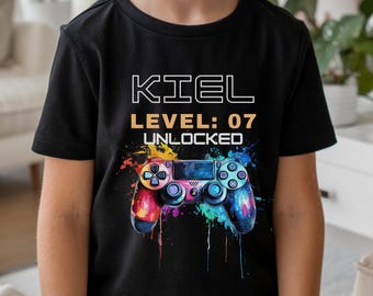 Könnte beinhalten: Schwarzes T-Shirt mit einem farbenfrohen Videospiel-Controller-Motiv. Das Shirt zeigt den Namen "KIEL", den Text "LEVEL: 07 UNLOCKED" und ein Controller-Design im Aquarellstil.