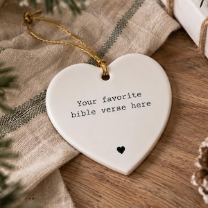Op de afbeelding: Een wit hartvormig keramisch ornament met de zwarte tekst "Your favorite bible verse here" en een klein zwart harticoontje aan de onderkant. Het ornament is voorzien van een goudkleurig koordje om het op te hangen aan een boom of standaard.