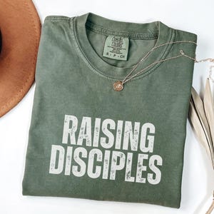 Könnte beinhalten: Ein grünes T-Shirt mit dem Aufdruck "Raising Disciples" in Weiß auf der Vorderseite. Das Shirt hat einen Used-Look und ist aus einem weichen, bequemen Stoff.