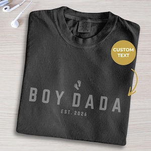 May include: Dark grey t-shirt featuring the text "BOY DADA" and "EST. 2026" in light grey, with baby footprints. The shirt is folded on a light wood surface.