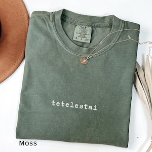 May include: A moss green Comfort Colors t-shirt with the word "tetelestai" printed in white. A gold necklace with a pendant rests on the shirt. The shirt is folded and displayed with a brown hat and decorative leaves.