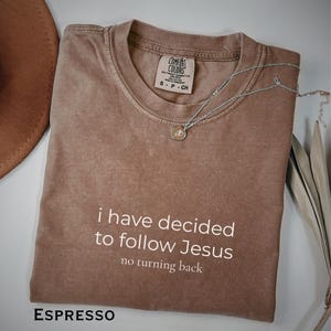 以下が含まれることがあります： 「i have decided to follow Jesus, no turning back」の白文字がプリントされたエスプレッソカラーのTシャツ。ペンダント付きのシルバーネックレスがシャツの上にあります。背景には茶色の帽子が見えます。