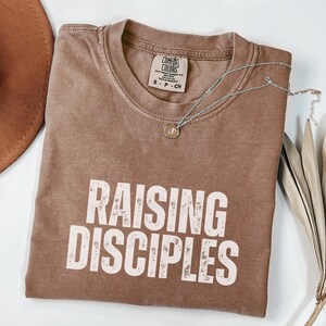May include: A brown t-shirt with the text "RAISING DISCIPLES" in white lettering. The shirt has a distressed look and is folded in half.