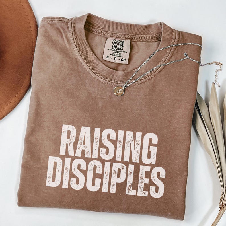 May include: A brown t-shirt with the text "RAISING DISCIPLES" in white lettering. The shirt has a distressed look and is folded in half.