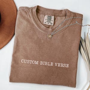 May include: A brown t-shirt with the text "CUSTOM BIBLE VERSE" printed on the front. The shirt is made of a soft, comfortable fabric and has a relaxed fit.