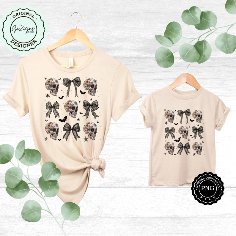 May include: Two beige t-shirts, one adult and one child size, displayed on wooden hangers. Each shirt features a repeating pattern of floral skull designs, black bows, bats, and stars. The shirts are labeled "Original Designer" and "Digital Download".