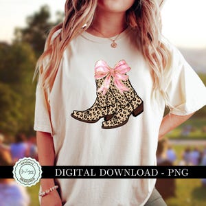 K&ouml;nnte beinhalten: Ein wei&szlig;es T-Shirt mit einem Design aus zwei Cowboy-Stiefeln im Leopardenmuster mit einer rosafarbenen Schleife oben. Der Text "DIGITAL DOWNLOAD - PNG" befindet sich am unteren Rand des Shirts.