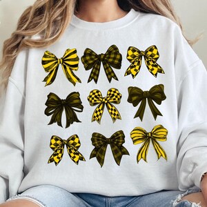 May include: White sweatshirt featuring a collection of nine bow illustrations. The bows vary in design, with patterns including yellow and black stripes, checkered patterns, and solid dark green. The bows are arranged in three rows of three.