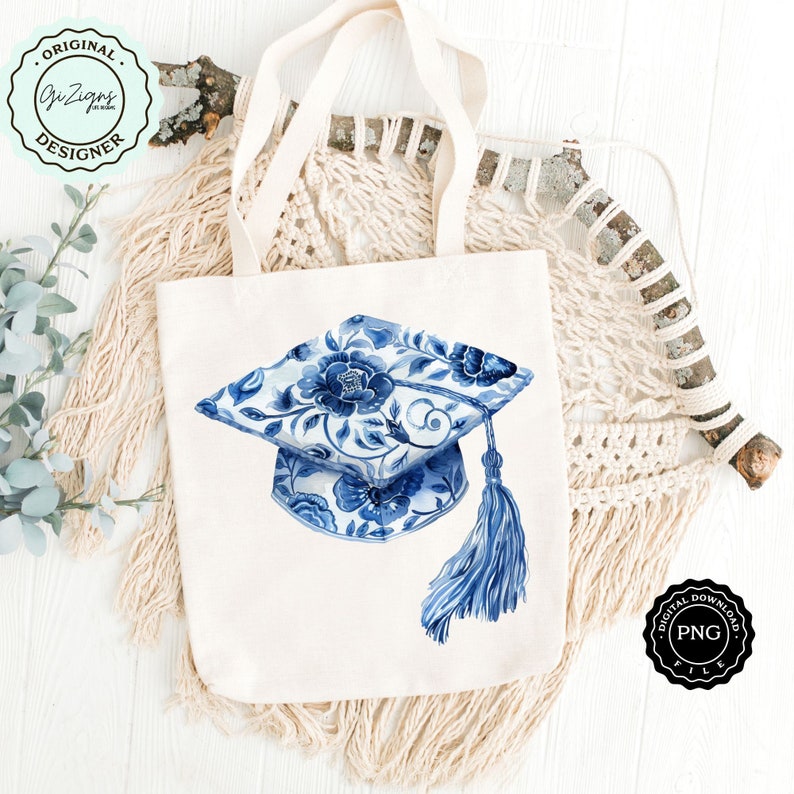 May include: A white tote bag with a blue and white floral graduation cap design. The bag is sitting on a white wooden surface with a macrame wall hanging in the background.
