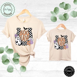 May include: Two beige t-shirts, one adult and one child size, with a Halloween-themed design. The design features a horseshoe with the words "BOO HAW", a ghost, flowers, and a bat. The horseshoe has a black and white checkered pattern.