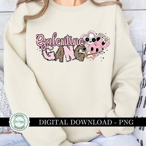 May include: A cream-colored sweatshirt with the text "Galentine Gang" in pink and black letters. The text is surrounded by pink hearts with black outlines and a pink and white checkered pattern.