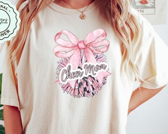 May include: Cream-colored t-shirt featuring a pink bow and a pom-pom design with the words "Cheer Mom" in a script font. The design is centered on the front of the shirt. The shirt has a relaxed fit.