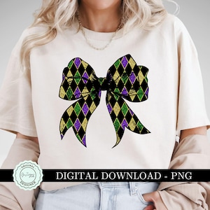 May include: A white t-shirt with a purple, green, and gold bow design. The bow is made of a diamond pattern and has a glittery texture. The text "Original Designer" is on a circle in the lower left corner.