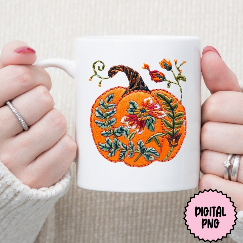 Peut inclure: Une tasse en c&eacute;ramique blanche avec un motif de citrouille brod&eacute; &agrave; la main. La citrouille est orange avec des feuilles vertes et des fleurs rouges. La tasse a une anse.