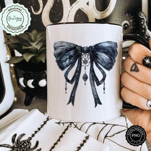 May include: White ceramic mug with a black rim and handle. The mug features a watercolor illustration of a large black bow with a silver and black pendant.
