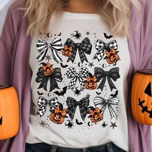 May include: Cream-colored t-shirt featuring a Halloween-themed design. The design includes various black and white bows, orange pumpkins with witch hats, spiders, bats, stars, and crescent moons. The shirt is worn by a person.
