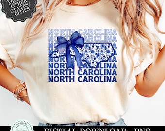 May include: A white t-shirt featuring a blue and white design of the state of North Carolina. The design includes a blue bow and floral accents. The words "North Carolina" are repeated in a stacked design.