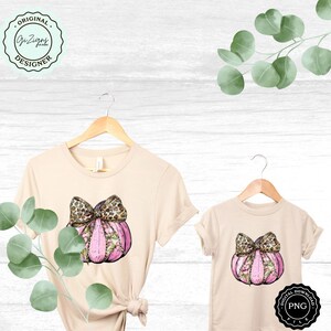 Pode incluir: Duas camisetas bege com uma ab&oacute;bora rosa com um la&ccedil;o de estampa de leopardo em cada uma. A ab&oacute;bora tem detalhes florais e &eacute; cercada por folhas verdes. As camisetas est&atilde;o penduradas em cabides.