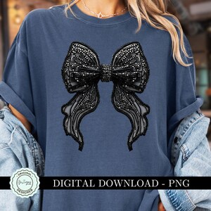 May include: A blue t-shirt with a black rhinestone bow design. The design is a large bow with a center knot and flowing tails. The shirt has a crew neck and short sleeves.  "Original Designer" is printed in a circle on the bottom left corner of the shirt.