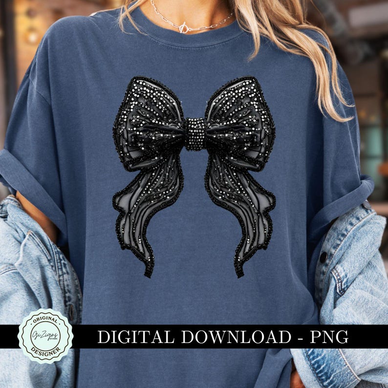 May include: A blue t-shirt with a black rhinestone bow design. The design is a large bow with a center knot and flowing tails. The shirt has a crew neck and short sleeves.  "Original Designer" is printed in a circle on the bottom left corner of the shirt.